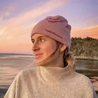 Frau mit Mütze lächelt zum Meer bei Sonnenuntergang, warm bekleidet. – Website Baukasten