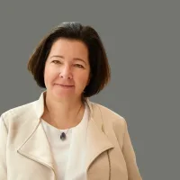 Frau mit braunen Haaren in weißer Jacke vor grauem Hintergrund.
